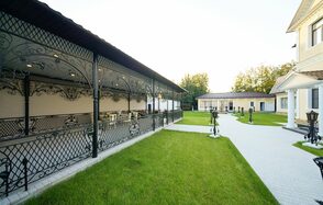Загородный дом. Открытая терраса с коваными декоративными элементами и навесом, столы и стулья, рядом выложенная плиткой дорожка, окруженная зелёным газоном, фонари у дорожки, светлое двухэтажное здание справа и одноэтажное впереди, деревья на заднем плане.