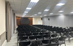 КПЦ Единство. Конференц-зал с рядами черных стульев перед экраном, натянутыми серыми шторами и колонками по бокам. Белый потолок с встраиваемыми светильниками. Светлые стены с акцентом из дерева вокруг экрана.