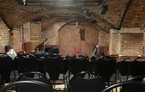 Культурный центр «Хитровка». "Арт-зал". Подвал с кирпичными стенами и арками, сценическое пространство с деревянным фоном и малой сценой, черные складные стулья для зрителей, потолочные светильники сценического освещения. Компактная, уютная атмосфера в полуподвальном помещении.