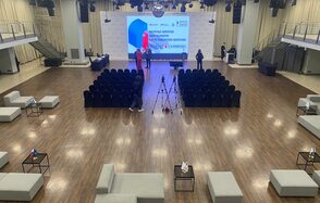 Зал «Азимут». Просторное помещение с деревянным полом, сцена с экраном на заднем плане, ряды черных стульев для зрителей, серые диваны и столы по периметру, светлая драпировка на стенах, подвесное освещение, камеры и микрофоны на триподах, лестницы по бокам.