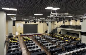 Зал «Азимут». Просторное конференц-пространство с рядами длинных столов и стульев, сцена с экраном. Черный натяжной потолок с подвесным освещением и звуковым оборудованием. Лампы квадратной формы, бежевые стены и шторы, деревянный пол, дополнительный балкон для зрителей.