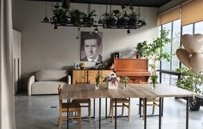 Пространство СемЬСюР с Кухней. Просторная комната с серой бетонной плиткой на полу. В центре деревянные столы со стульями, вазы с цветами. На заднем плане пианино, диван, шкаф. Светло-серые стены, декорированы фотографией. Большие окна с жалюзи, потолочные светильники. Комнатные растения висят и стоят вдоль стен.