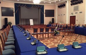 Концертный зал Ассамблея. Просторный зал с деревянным паркетным полом, столы и стулья с синими скатертями и обивкой, множество микрофонов на столах. На сцене экран с занавесом, рядом флипчарт. Освещение представлено люстрой и точечными светильниками.