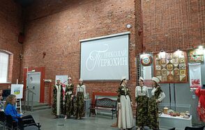 Выставочный зал Форум. Кирпичное помещение с высоким потолком. На стене проекционный экран с надписью. Слева металлические двери, справа экспонаты и яркие ткани. Несколько людей в традиционных костюмах. В зале стулья для зрителей.