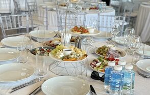 Event  Hall ARCOBALENO (Эвент Холл АРКОБАЛЕНО) . Круглый белоснежный стол, сервированный на банкет. Прозрачные стулья, белая посуда, бокалы, столовые приборы. Центр украшен закусками на металлической подставке. На столе бутылированная вода, овощи и фрукты, кодовые знаки отсутствуют.