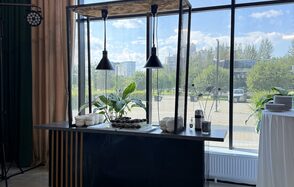 Event  Hall ARCOBALENO (Эвент Холл АРКОБАЛЕНО) . Современный интерьер с барной стойкой и подвесными черными светильниками у панорамного окна, металлическая конструкция с растениями, стеклянные стены, бежевые шторы, белая скатерть на круглом столе с тарелками.