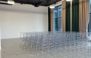 Event  Hall ARCOBALENO (Эвент Холл АРКОБАЛЕНО) . Зал с высокими потолками и большими окнами, занавешенными зелеными и бежевыми шторами. Ряды прозрачных стульев расставлены на сером плиточном полу. Две круглые подвесные лампы на черном потолке. Белая стена без украшений.