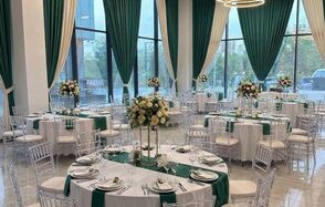 Event  Hall ARCOBALENO (Эвент Холл АРКОБАЛЕНО) . Банкетный зал с круглыми столами, украшенными белыми скатертями и зелеными дорожками. Прозрачные стулья, большие окна с зелеными шторами. Центральные композиции из светлых цветов. Потолочное освещение в виде кольцевых светильников.