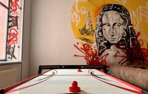 Арт Хаус. Комната с настольным аэрохоккеем и граффити на стене, изображающим портрет. Открытое окно слева, бежевый диван справа. Стены белые с яркими граффити в красных, черных и желтых цветах.