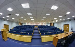 Конференц-зал Петровский. Аудиторное помещение с рядами синих кресел и деревянными столами. Спереди кафедра с трибуной и микрофонами. Подвесной потолок с встраиваемыми светильниками. Пол синий ковровый. Окна закрыты жалюзи. Две колонки по бокам.
