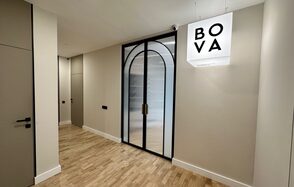 Лекторий на 53 этаже Москва-Сити - 100 м², до 35 человек. Коридор с деревянным полом, светлыми стенами, и черной стеклянной дверью с арочной формой. На стене справа белый светильник подвесной с четырьмя буквами: BOVA. Две двери видны сбоку и в конце коридора.
