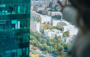 Лекторий на 53 этаже Москва-Сити - 100 м², до 35 человек. Высотное здание с зеркальными окнами, через которые видны офисные помещения. Вид на город с множеством жилых зданий, разной этажности. Внизу зелёные деревья и дорога. Размытая рука на переднем плане указывает на вид из окна.