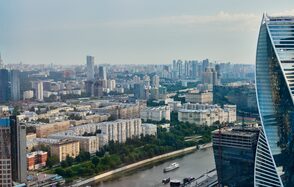 Квартира площадью 60 м² для мероприятий Свидания в Москва Сити. Вид на городскую панораму с высоты, река с лодкой, многоэтажные жилые здания разного дизайна, открытые зелёные пространства, современное стеклянное здание справа, светлое небо.