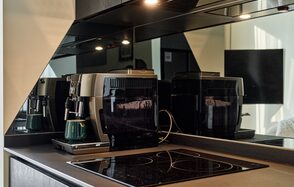 Квартира площадью 60 м² для мероприятий Свидания в Москва Сити. Кухонное пространство с кофемашиной и варочной панелью на темной столешнице. Зеркальный фартук, черные кухонные шкафы. Встроенное освещение в нижней части шкафов. Светлые стены, отсутствующий текст.