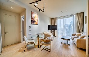 Квартира площадью 60 м² для мероприятий Свидания в Москва Сити. Современная гостиная с деревянным полом, большими окнами и светлыми шторами. Мебель включает два дивана, стол со стеклянной столешницей и стулья. На стене картина, на потолке чёрные светильники. Есть телевизор на стойке.