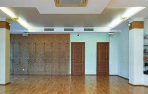 Спортзал Йога-студия площадью 100 м². Просторное помещение с деревянным полом, светлыми стенами и натяжным потолком. На одной из стен канаты и две двери. Система освещения встроена в потолок. На другой стене открытые стеллажи. Вентиляционные решетки под потолком.