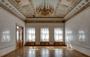 Особняк Сухозанета. Просторное помещение с паркетным полом, тремя большими окнами с белыми шторами, резными наличниками. Стены светлые, с лепниной. Потолок украшен росписью и массивной люстрой. Деревянные двойные двери, классический интерьер.