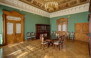Особняк Сухозанета. Просторная комната с высокими потолками и паркетным полом, стены окрашены в зеленый цвет. Деревянные двери и мебель с классическими элементами. Пара стульев вдоль стен, круглый стол с мягкими стульями в центре. Люстра и обильный декор на потолке.