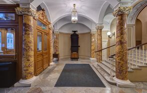 Особняк Сухозанета. Вестибюль с арочными сводами и мраморными колоннами, деревянная дверь слева, центральный ковер на плиточном полу, лестница с деревянными перилами справа, висячая люстра и настенные светильники, преобладающие бежевые и коричневые тона.