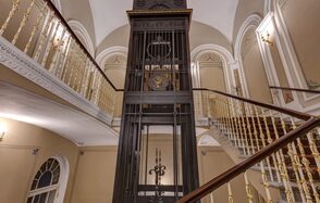 Особняк Сухозанета. Винтовая лестница с золотистыми перилами, центральный лифт в металлической шахте черного цвета, стены бежевые с белой лепниной, декоративные светильники, арочные окна, мраморные и деревянные элементы, высокие потолки.
