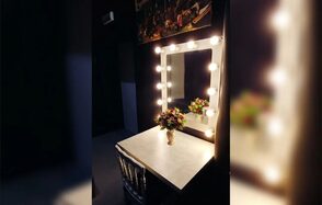 ДАЙТЕШУМА.РФ. Гримерное пространство с зеркалом в белой раме, окруженном лампочками. Белый стол под зеркалом и стул с прозрачной спинкой. Ваза с цветами на столе. Темные стены, небольшой верхний светильник на стене.