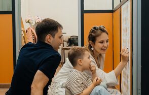 Дом Жар-Птицы. Закрытое пространство с оранжевыми и серыми перегородками. Деревянный пол, белые стены. На заднем плане стол с бумагами и аксессуары. Женщина, мужчина и ребенок, сидящие на полу, смотрят на стену с рисунками.