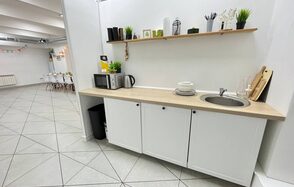 Зал площадью  122 м 2. Белая кухня с деревянной столешницей, раковина, микроволновка, чайник. На полке кухонные принадлежности и декор. Белые стены, серый плиточный пол. В соседней комнате столы и стулья в светлой гамме, на стене гирлянда.