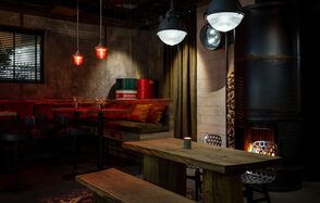 Kano loft. Красивое пространство с деревянной мебелью, длинными скамьями и столом. Подвесные светильники с теплым красным и белым светом. Стена из серого бетона, металлическая печь, металлические бочки. Тёмные шторы, ковёр с геометрическим рисунком.