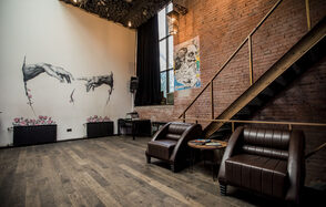 Street Art Loft (Стрит Арт Лофт). Лофт-пространство с кирпичными стенами, деревянными полами, лестницей и высокими потолками. На светлой стене граффити с пятью руками и розовыми цветами. Два кожаных кресла и круглый столик. Большое окно за закрытой шторой.