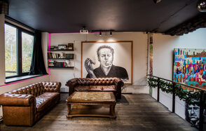 Street Art Loft (Стрит Арт Лофт). Лофт-пространство с деревянным полом, черными потолками, большими окнами, двумя коричневыми кожаными диванами, деревянным столом, настенной живописью, книжными полками и произведением искусства с изображением курящего человека.