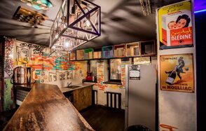 Street Art Loft (Стрит Арт Лофт). Барное пространство с деревянной стойкой, стеллажами с книгами и плакатами на стенах. Потолочные светильники в металлической раме. Стены с граффити и яркими акцентами. Холодильник серого цвета. Тёмный пол, подчеркнутый освещением.