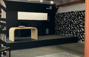 Бертгольд центр. Просторный лофт. Сценическая площадка с деревянным столом, видеопроектором и экраном. Стена с черно-белым графическим узором. Потолок с открытыми коммуникациями, освещение из точечных светильников. Пол темно-серый, напротив сцены ковер светлого цвета.