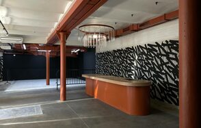 Бертгольд центр. Просторный лофт. Просторное помещение с бетонными полами, открытыми балками и колоннами кирпичного цвета. Темные стены с графическим черно-белым узором, округлый деревянный стол. Подвесное освещение с прозрачными элементами, кондиционер на потолке.