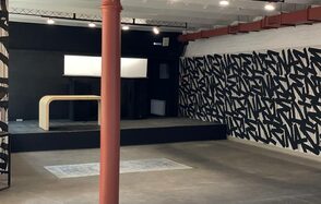 Бертгольд центр. Просторный лофт. Арендное пространство в индустриальном стиле с бетонным полом и железной колонной, черной сценой, светлым деревянным столом, черными и белыми стенами с графическим рисунком, светильниками на потолке и белым светодиодным экраном на сцене.