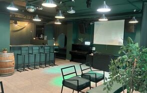 Work Shop Loft(Воркшоп Лофт). Зал 11 . Открытое помещение с барной стойкой и высокими стульями, фортепиано, экран для проектора, металлические подвесные светильники, деревянная бочка, черные стулья с мягкими подушками, зеленые стены и потолок, комнатное растение, бежевый пол.