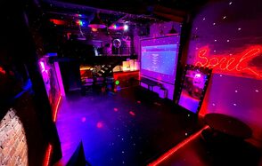 Soul Loft . Тускло освещенное пространство с разноцветными огнями, баром и большим экраном. На стене написано слово Soul в ярко-красных огнях, что создает живую атмосферу, подходящую для встреч или мероприятий. Деревянный пол добавляет теплоты современному декору.