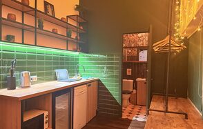 Barshowplace (БарШоуПлейс). Лофт для вечеринок с коктейльным баром . Кухонное пространство с деревянной мебелью и барной стойкой, зелёная плитка на стенах, открытые полки с посудой и декором. Освещение желтоватого оттенка. Видна дверь в санузел, частично облицованный плиткой с узором. Напольное покрытие из тёмного дерева.