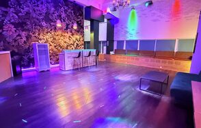 Barshowplace (БарШоуПлейс). Лофт для вечеринок с коктейльным баром . Просторное помещение с барной стойкой и стульями, диванами у стены. Темный деревянный пол, цветное освещение, декоративные обои с растительным рисунком. Белая кирпичная стена, разноцветные световые проекции. Люстра на потолке.