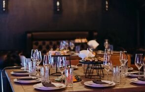 Двухуровневый лофт-бар с потолками 5 м. Ресторан, сервированный стол для большой компании, темные стены, мягкая кожаная мебель, стеклянные бокалы, тарелки с темными салфетками, металлические подставки, приглушенное освещение, свечи.