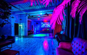 Лофт Комод. Уютный лофт со сценой, караоке и барбекю. Место для вечеринок с диванами и столиками вдоль стен. Центральное пространство свободно, на стене большой экран. Потолочные светильники, разноцветное освещение в синих и фиолетовых тонах. Пол из светлого ламината, декор из растений.