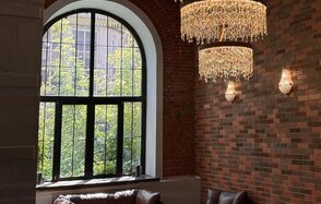 Brick loft with large windows. Арочное окно с черной рамой, кирпичная стена разных оттенков, два хрустальных светильника, кожаные кресла коричневого цвета, дневной свет, две декоративные свечи на подоконнике.