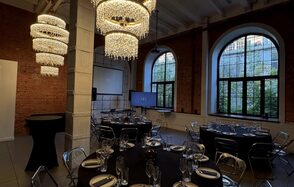 Brick loft with large windows. Уютное праздничное пространство с круглыми столами, черными скатертями, прозрачными стульями. Декорировано массивными люстрами. Красные кирпичные стены с большими арочными окнами. Экран установлен у стены. Белые тарелки и бокалы расставлены на столах.