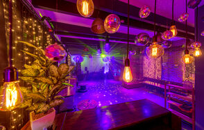 Для вечеринок с супер звуком. Праздничное пространство с деревянными столами и лавками, освещение из подвесных ламп и диско-шаров, яркие цветные прожекторы, светодиодные гирлянды, живая зелень в горшках, кирпичные стены, деревянные балки, фиолетовая и оранжевая цветовая гамма.