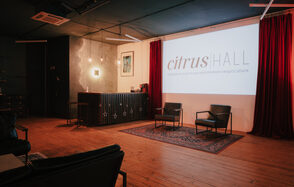 Citrus Hall (Цитрус Холл). Лофт Грейпфрут. Пространство с деревянным полом и тёмным потолком. Экран с надписью, два кресла на ковре, барная стойка с декоративной подсветкой. Красные шторы, подвесные светильники, картина на стене. Интерьер в тёмно-красных и чёрных оттенках.