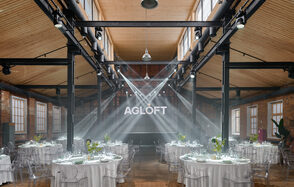 AG Loft. Urban (АГ Лофт. Урбан). Просторный лофт с раритетным автомобилем. Просторное лофт-пространство с деревянным потолком и кирпичными стенами. На полу деревянное покрытие. Расставлены круглые столы с белыми скатертями и прозрачными стульями. Яркое освещение сверху. На задней стене светящийся знак "AGLOFT". Световые прожекторы направлены к стене.