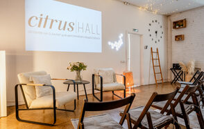 Citrus Hall (Цитрус Холл). Зал Свити. Просторное помещение с деревянным полом, белыми кирпичными стенами и проектором. Есть стулья, кресла и круглый стол с вазой и цветами. Декоративные элементы: лестница, настенные часы, световые гирлянды и крупный текст проекта.
