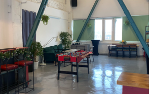 Work Shop Loft(Воркшоп Лофт). Зал 2 с верандой. Лофт-пространство с высокими потолками и скошенной крышей. Центр оборудован настольным футбольным столом. Мягкая мебель: диван и стулья. Есть длинный стол с черными стульями. Зелёные растения, светло-серые стены, большие окна, блестящий пол.