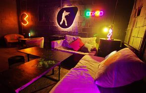 Лофт Геймеры. Крупный план диванов, между ними стоит бочка. Рядом расположен стол, вдалеке стоят кресла и ещё один стол. На стене неоновые лампы и игровые логотипы