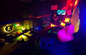 Лофт Геймеры. Второй этаж лофта. К стене прикреплены небольшие картины с телевизором. На полу стоит небольшой столик с пуфами, в углу маленькая тумбочка с лампой. Также видно убранство первого этажа