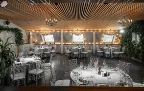 Loft - bar under closure in Peterhof. Банкетный зал с деревянным потолком и люстрами, круглые столы накрыты белыми скатертями, прозрачные стулья, настенные окна с занавесками и световой гирляндой, по бокам зелёные растения, тёмный пол, сервировка с бокалами и приборами.